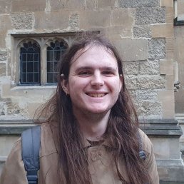 The photograph shows a young white man, with long brown hair, standing in front of a stone wall. He is wearing a light brown jacket, and is smiling.
