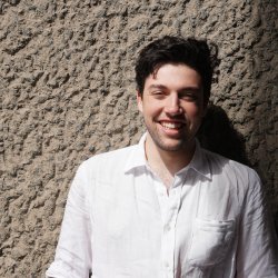 A Picture of Ned smiling in bright sunshine and a white shirt in front of a wall.