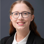 A portrait of Georgina. She is a woman with ginger hair, white skin, and blue glasses. Georgina is smiling and wearing a white collared shirt and black blazer. 