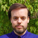 Portrait of Dan Hill, wearing a blue button-up shirt and standing in front of a bush (out of focus)