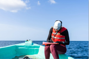 A researcher sits on a dinghy at sea, writing on a clipboard