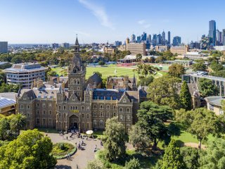 Photo from above of the Uni of Melbourne, an old grand building surrounded by trees and playing fields, with city skyscrapers in the background Photo from above of the Uni of Melbourne, an old grand building surrounded by trees and playing fields, with city skyscrapers in the background