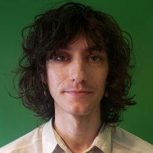 A man with brown hair and a white shirt, stood in front of a green background.